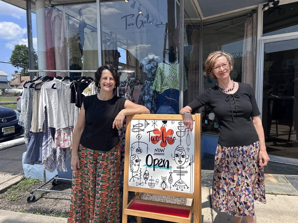 The two gals outside their vintage shop with a colorful Now Open sign and a rack of vintage clothes